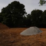 A cleared dirt area with a pile of gravel and a pile of soil, likely prepped for building or excavating a pole barn, surrounded by tall trees under a cloudy sky. Kentucky Builders And Excavating