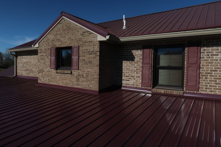 A house with brick walls, maroon shutters, and a shiny maroon metal roof stands under a clear blue sky. Sunlight creates strong shadows on the building and roof, highlighting its classic charm. Kentucky Builders And Excavating