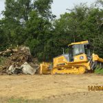 A yellow bulldozer pushes a pile of debris and tree roots on a dirt clearing near a wooded area, excavating the site for a new building. The scene is outdoors, dated 14/09/2020 at 03:04. Kentucky Builders And Excavating