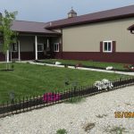 A modern beige and maroon pole barn building with a covered entrance, small garden beds, flowers, a young tree, and neatly trimmed grass. The photo was taken on September 16, 2020, at 2:44. Kentucky Builders And Excavating