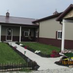 A modern pole barn-style house with beige and burgundy siding, a covered porch, manicured lawn, flower beds, and a wheelbarrow near the sidewalk. The date 16/09/2020 is displayed on the photo. Kentucky Builders And Excavating