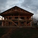 A large wooden pole barn frame under construction stands against a cloudy evening sky, surrounded by bare trees and grass, with exposed beams and scaffolding visible—evidence of active building on the site. Kentucky Builders And Excavating