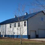 A large white and blue pole barn with two garage doors and a metal roof sits on a gravel driveway, surrounded by bare trees and fields under a clear blue sky. Kentucky Builders And Excavating