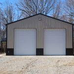 A metal pole barn with two large white garage doors stands on a gravel driveway, surrounded by leafless trees and bare ground on a clear day—ideal for building projects or excavating work. Kentucky Builders And Excavating