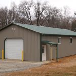 A tan metal pole barn with a green roof and four windows sits on a cleared lot with gravel and dirt, surrounded by trees and grassy areas in the background. Kentucky Builders And Excavating