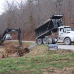 A dump truck unloads gravel onto a rural road while a yellow excavator works near a drainage pipe, excavating for a future building. Leafless trees and a construction sign are visible in the background. Kentucky Builders And Excavating
