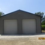 A gray metal pole barn with two large white garage doors stands on a concrete driveway, surrounded by trees and grass. The photo is dated 12/03/2021 in the bottom right corner. Kentucky Builders And Excavating
