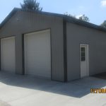 A large gray metal pole barn with two closed white roll-up doors and a single entry door sits on a concrete driveway, surrounded by grass and trees under a blue sky. Kentucky Builders And Excavating