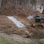 A small tracked loader spreads rocks over a dirt slope for building a pole barn, while another excavator works in the background, surrounded by bare trees and earth. Kentucky Builders And Excavating