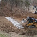 A compact track loader spreads white gravel on a dirt path in a clearing, while an excavator works on excavating near a forested slope, preparing the site for building. Kentucky Builders And Excavating