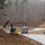 Construction vehicles, including an excavator, dump truck, and bulldozer, work on grading and laying gravel on a rural road near a pole barn, surrounded by bare trees and a hilly landscape. Kentucky Builders And Excavating