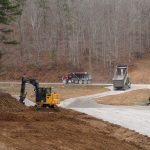 Construction vehicles, including an excavator, dump truck, and bulldozer, are building a rural road surrounded by leafless trees in late autumn or winter. Gravel is being spread as part of the work near a future pole barn site. Kentucky Builders And Excavating