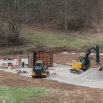 Two yellow construction vehicles are excavating a gravel area at a building site. Several workers stand near equipment and materials, with a metal pole barn structure and forested hills in the background. Kentucky Builders And Excavating