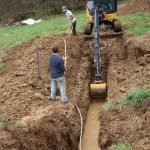 A small excavator digs a muddy trench for a pole barn as two people stand nearby, measuring its width with a tape measure in a grassy, rural area. Kentucky Builders And Excavating
