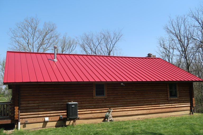 A log cabin building with a bright red metal roof sits on green grass. Leafless trees stand in the background, and an air conditioning unit is near the wall. The sky is clear and blue. Kentucky Builders And Excavating
