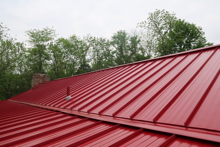 A bright red metal roof with raised seams tops this building, set against trees and a cloudy sky. A small chimney and a vent are visible on the roof. Kentucky Builders And Excavating