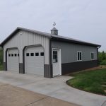 A gray metal pole barn building with two white garage doors, a white entry door, and a cupola on the roof sits beside a concrete driveway, surrounded by grass and trees—the result of expert excavating and thoughtful design. Kentucky Builders And Excavating