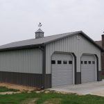 A gray metal two-car garage, built in a classic pole barn style with double doors and a small cupola on the roof, is attached to the side of a brick house and sits on a concrete driveway next to a grassy lawn. Kentucky Builders And Excavating