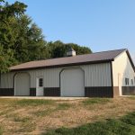 A large, metal-sided pole barn with two white garage doors, a white entry door, and a brown roof sits on a grassy lot beside tall green trees and freshly excavated ground under a clear blue sky. Kentucky Builders And Excavating