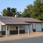 A modern, single-story pole barn with two large garage doors, a covered entrance, and light brown siding sits on a concrete lot surrounded by green trees. Kentucky Builders And Excavating