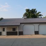 A metal pole barn with a slanted roof, two white garage doors, and a single entry door sits on a concrete driveway, surrounded by green trees and grass in the background. Kentucky Builders And Excavating