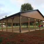 A large, open-sided pole barn with a metal roof and wooden supports stands on a concrete slab in a grassy park area, with trees and buildings visible in the background. Kentucky Builders And Excavating