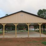 A large, open-sided pole barn with a metal roof and wooden supports stands on a concrete slab, surrounded by green grass and trees in an outdoor park setting. Kentucky Builders And Excavating