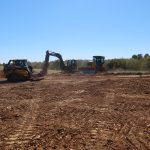 Three large construction vehicles, including a bulldozer, excavator, and loader, are excavating a dirt construction site under a clear blue sky, with trees and mounds of earth in the background. Kentucky Builders And Excavating