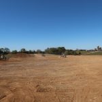 Construction site with cleared dirt ground, piles of soil, and construction vehicles excavating under a clear blue sky. Trees and a few buildings, possibly including a pole barn, are visible in the distant background. Kentucky Builders And Excavating