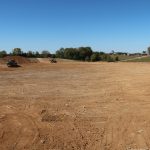A wide, freshly graded dirt field ready for building a pole barn, with construction vehicles and piles of soil in the distance, surrounded by green trees under a clear blue sky. Kentucky Builders And Excavating