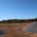 Large mounds of gravel and dirt sit on a cleared construction site, ready for excavating a new building, with trees visible in the background under a clear blue sky. Kentucky Builders And Excavating