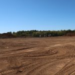 A wide, flat construction site with dirt, tire tracks, and scattered mounds of soil under a clear blue sky—perfect for excavating or preparing to build a pole barn. Trees and construction vehicles are visible in the distant background. Kentucky Builders And Excavating