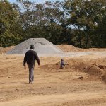 An adult walks toward a small child who is playing in a sandy construction area, where piles of dirt and gravel hint at recent excavating, surrounded by trees in the background under a clear sky. Kentucky Builders And Excavating