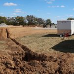 A trench dug through a grassy field on a construction site for a pole barn, with piles of dirt, a white utility trailer, and equipment under a clear blue sky. Trees and gravel piles are in the background. Kentucky Builders And Excavating