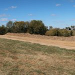 A dirt road runs through a grassy field toward a distant farm with silos, trees, and a pole barn near a pile of gravel under a blue sky with scattered clouds. Kentucky Builders And Excavating