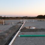 A construction site at sunset with exposed pipes laid in trenches across sandy ground, surrounded by dirt piles and excavating equipment. Trees and distant hills are visible in the background. Kentucky Builders And Excavating
