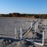 A construction site excavating sandy soil for a pole barn, with exposed white PVC pipes, a person working in the background, construction vehicles, and dirt piles under a clear blue sky. Kentucky Builders And Excavating