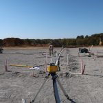 A construction site with surveying equipment on a tripod in the foreground, marked stakes in the ground, and workers and excavators excavating for a new building under a clear blue sky. Kentucky Builders And Excavating