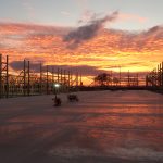 Construction site at sunset, featuring wooden framing for a pole barn on both sides, a smooth concrete floor, scattered equipment, and a dramatic orange and pink sky with clouds in the background. Kentucky Builders And Excavating