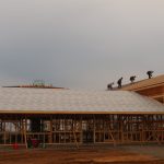 Several construction workers are building the roof of a large wooden pole barn frame at a construction site, with an overcast sky in the background. Kentucky Builders And Excavating