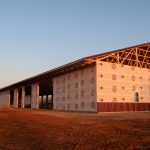 A large pole barn under construction, covered in white weatherproof sheeting, sits on reddish dirt ground in the golden light of sunset. Part of the roof framing is exposed, with workers and some excavating activity visible near the structure. Kentucky Builders And Excavating