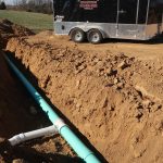 A trench with newly installed green PVC pipes for plumbing or drainage runs through dirt beside a silver trailer, highlighting the early stages of excavating and building a pole barn on the construction site. Kentucky Builders And Excavating
