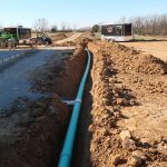 A long green pipe is laid in a trench at an excavating site, with dirt piled on both sides. Construction vehicles and trailers are visible in the background, along with a clear view of a pole barn on a sunny day. Kentucky Builders And Excavating