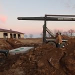 Two workers install a large concrete septic tank at a construction site, surrounded by dirt piles and excavating machinery, with a pole barn visible in the background as the sun sets. Kentucky Builders And Excavating