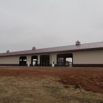 A large beige metal pole barn with a maroon roof stands under a cloudy sky, surrounded by bare earth—evidence of recent excavating—and patches of grass. The building features multiple wide openings and small cupolas on the roof. Kentucky Builders And Excavating