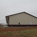 A large beige and brown metal pole barn stands on bare dirt and patchy grass under a cloudy, overcast sky. The side of the building has two small windows and no visible doors, hinting at recent excavating work. Kentucky Builders And Excavating