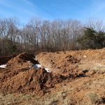 A large trench for a pole barn, with piles of brown soil on either side, sits in a grassy, open area near leafless trees under a clear blue sky. Small patches of snow are visible on the ground from recent excavating. Kentucky Builders And Excavating