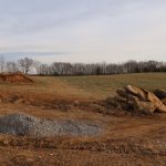 A grassy field with scattered trees in the background, piles of rocks, gravel, and dirt in the foreground suggest recent excavating for a pole barn or building, with tire tracks visible on the bare soil. Kentucky Builders And Excavating