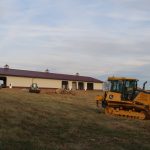 Construction vehicles, including a bulldozer and small excavator, are excavating a grassy slope in front of a large beige pole barn with a red roof under a partly cloudy sky. Kentucky Builders And Excavating