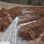 Trenches dug in a grassy field, possibly for building or excavating a pole barn, contain green pipes and white gravel, likely part of a septic system or drainage installation. Piles of dirt line both sides of the exposed trenches. Kentucky Builders And Excavating